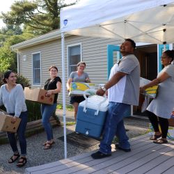 Five people smile and laugh as they bring boxes and a cooler out of a tan building